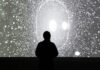 A silhouette of a person stands in front of a bright, abstract, starry or cosmic background projected on a large screen, creating a mysterious and contemplative scene.