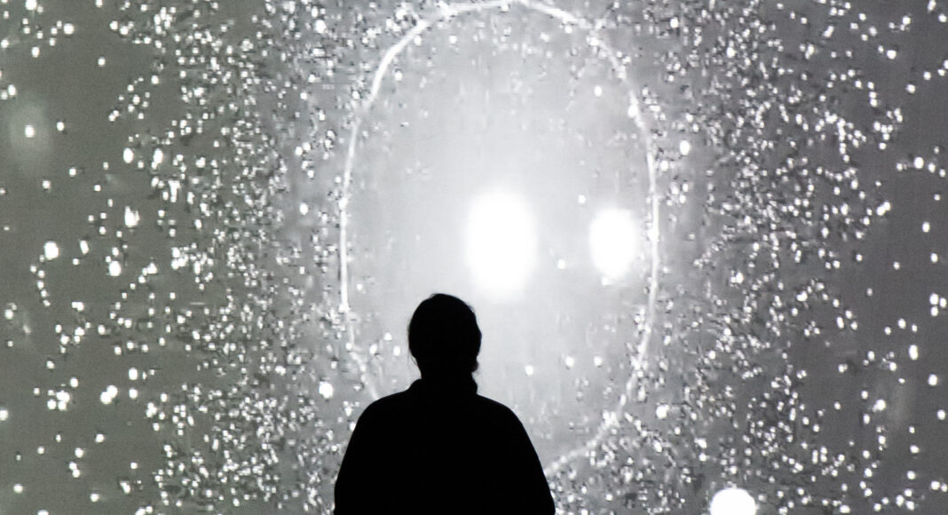 A silhouette of a person stands in front of a bright, abstract, starry or cosmic background projected on a large screen, creating a mysterious and contemplative scene.
