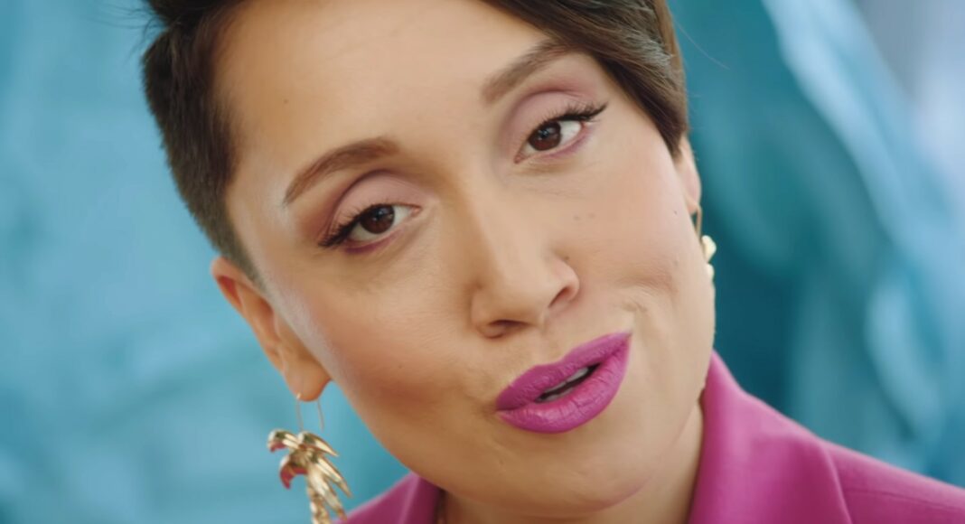 A person with short dark hair and pink lipstick wears a pink blazer and gold earrings, looking directly at the camera against a blue background.