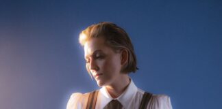 A person with short, light brown hair is wearing a white shirt, brown tie, and suspenders, looking down thoughtfully against a blue background with soft, warm lighting.