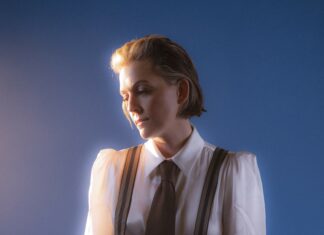 A person with short, light brown hair is wearing a white shirt, brown tie, and suspenders, looking down thoughtfully against a blue background with soft, warm lighting.