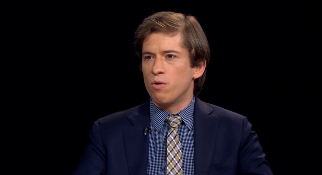 A man with light brown hair wearing a navy suit, blue checkered shirt, and plaid tie is speaking while seated against a plain black background.