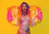 A person with colorful long hair and bold makeup poses confidently against a yellow background, wearing a sparkling bodysuit and dramatic fringed sleeves in shades of yellow, pink, and orange.