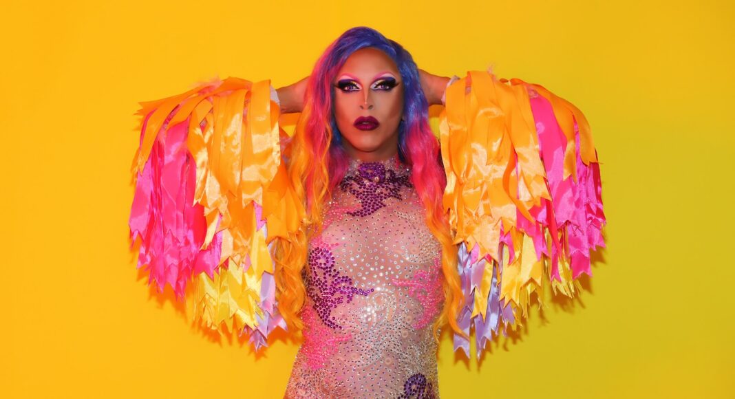 A person with colorful long hair and bold makeup poses confidently against a yellow background, wearing a sparkling bodysuit and dramatic fringed sleeves in shades of yellow, pink, and orange.