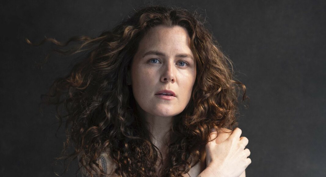 A woman with long, curly brown hair and floral tattoos on her arm gazes at the camera, holding her shoulder with one hand against a dark background.
