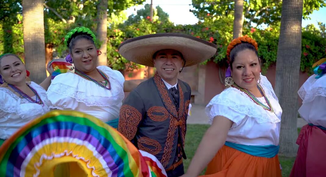 Four people in traditional Mexican attire perform outdoors. Three women wear colorful skirts and white blouses, and one man wears a charro suit and sombrero. They smile and dance amid green trees and sunlight.