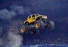 A yellow monster truck with BroDozer written on the side leaps into the air above a dirt arena, surrounded by tire smoke, with a large crowd watching in the stadium background.