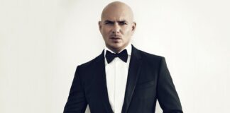 A bald man wearing a black tuxedo and bow tie stands against a plain light background, looking directly at the camera while buttoning his jacket.