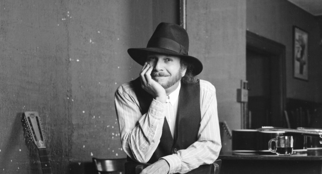 A man wearing a wide-brimmed hat, vest, and striped shirt sits backward on a wooden chair, resting his head on his hand and smiling. A guitar leans against the wall beside him, and a table with a glass and another guitar is nearby.