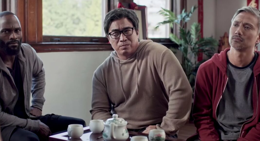 Three men sit together indoors, looking attentively toward someone out of frame. They are near a wooden table with tea cups and a teapot. The room has large windows and plants, creating a calm atmosphere.