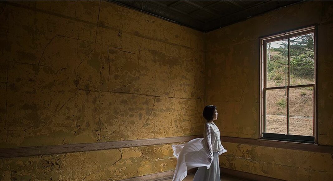 A woman in a white blouse and gray skirt stands in an empty, weathered room with peeling yellow walls, gazing out a window. Her blouse billows behind her as though caught by a breeze.