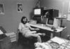 A man with long hair and a beard sits in a cluttered office, smiling near vintage computer equipment with monitors and papers. The workspace has scattered items, posters on the wall, and a cup on the desk.