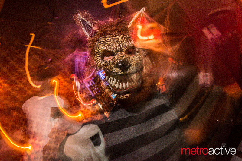 A person wearing a snarling werewolf mask and a striped shirt stands in a dark, colorful, and blurred setting; the image has vivid light streaks and the metroactive logo in the corner.
