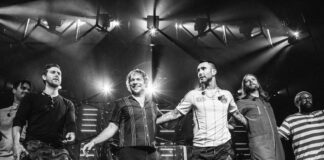 Six band members stand together on stage, holding hands and smiling under bright stage lights, with a drum set and concert equipment in the background. The image is in black and white.
