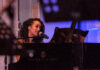 A woman with curly hair sings into a microphone while playing a black Steinway & Sons grand piano, bathed in soft purple lighting.