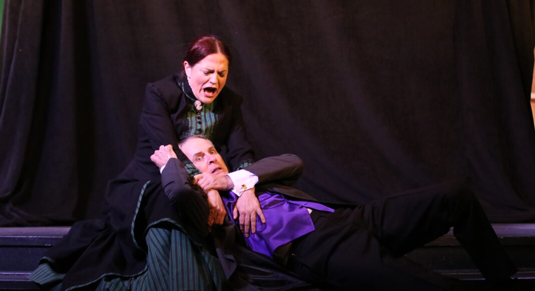 A woman in period costume holds a man in a suit and purple vest on the ground, gripping his face and neck, both with intense expressions, in front of a dark curtain on stage.