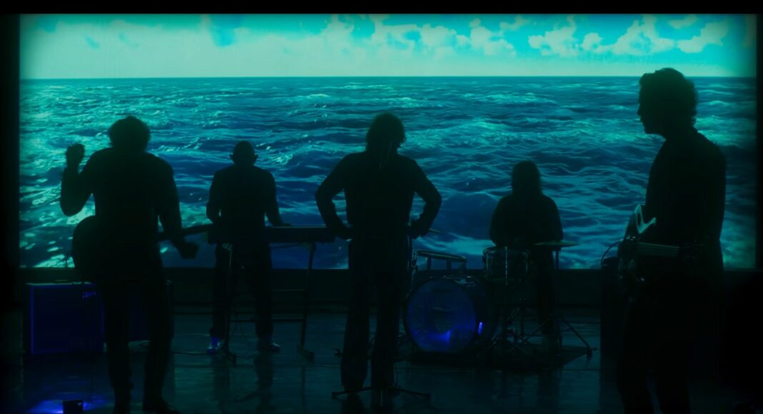 Five musicians stand in silhouette on stage with instruments, performing in front of a large screen displaying blue ocean waves under a cloudy sky.