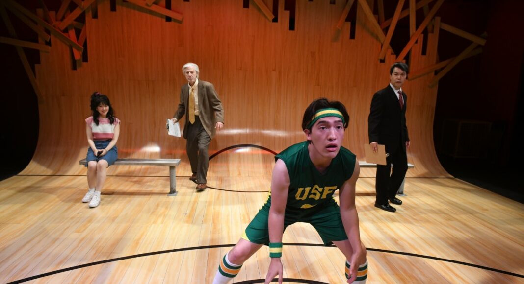 A young man in a green basketball uniform crouches on a gym floor set. Behind him, a girl sits on a bench, while two men—one older holding papers, one younger in a suit—stand in the background under dramatic lighting.