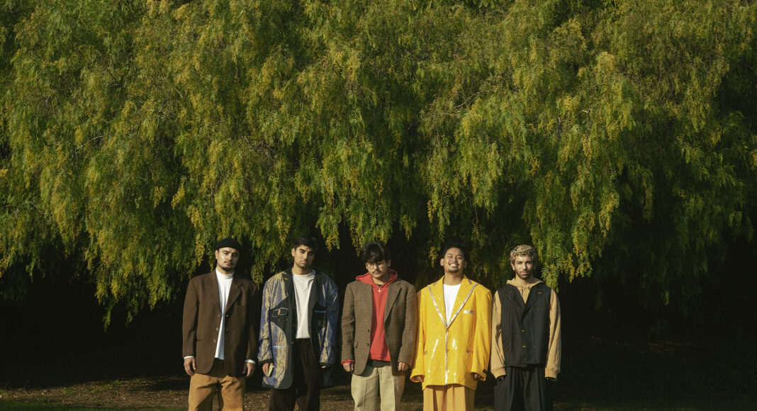 Five people stand in a row on grass in front of a large tree, each wearing colorful, oversized suits. The background is lush and green, suggesting a park or natural setting.