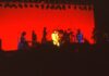 A band performs on stage under dramatic red lighting, with several musicians playing instruments and microphones set up, silhouetted against a brightly lit red background.