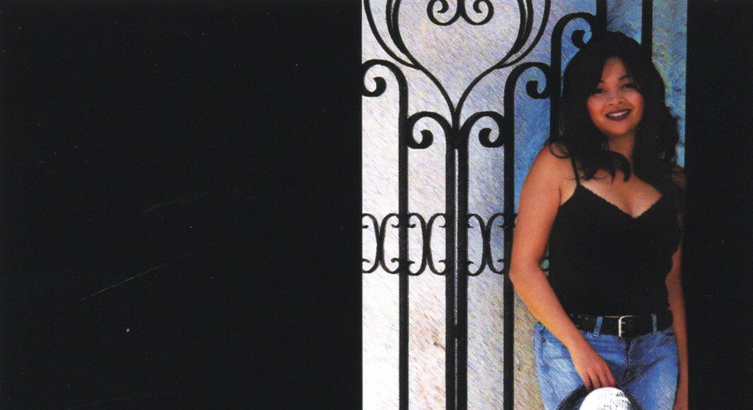 A woman in a black top and blue jeans stands by a wrought iron gate, smiling and holding a silver hat at her side. The left half of the image is mostly black.