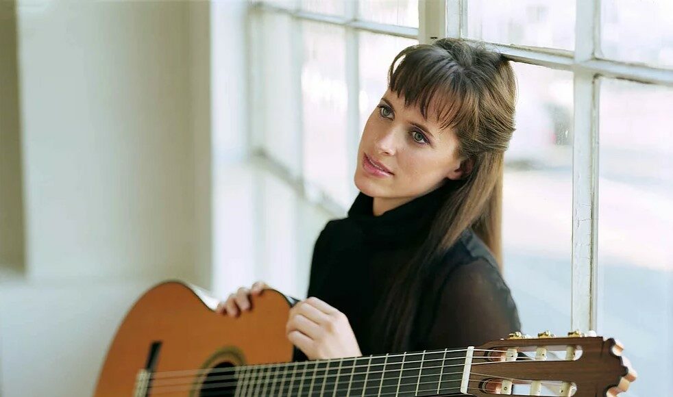 A woman with long brown hair, wearing a black top, sits by a large window holding a classical guitar. She looks thoughtfully at the camera, with natural light illuminating the scene.