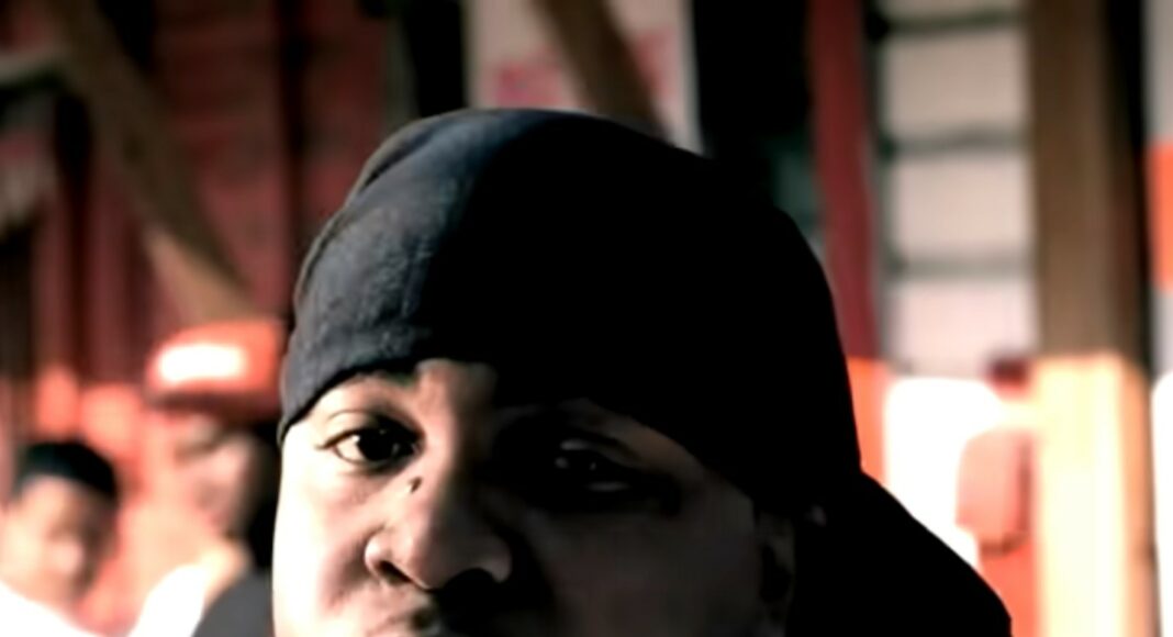 A man wearing a black durag and white shirt looks toward the camera, with blurred people and a building structure in the background.