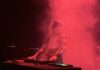 A person wearing a futuristic space suit and helmet stands behind a keyboard, performing on stage surrounded by dramatic red smoke.