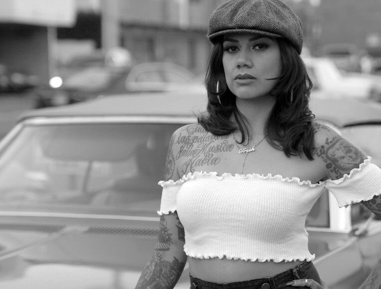 A woman with tattoos, wearing a newsboy cap and off-the-shoulder crop top, stands confidently in front of a classic car on a city street. The image is in black and white.