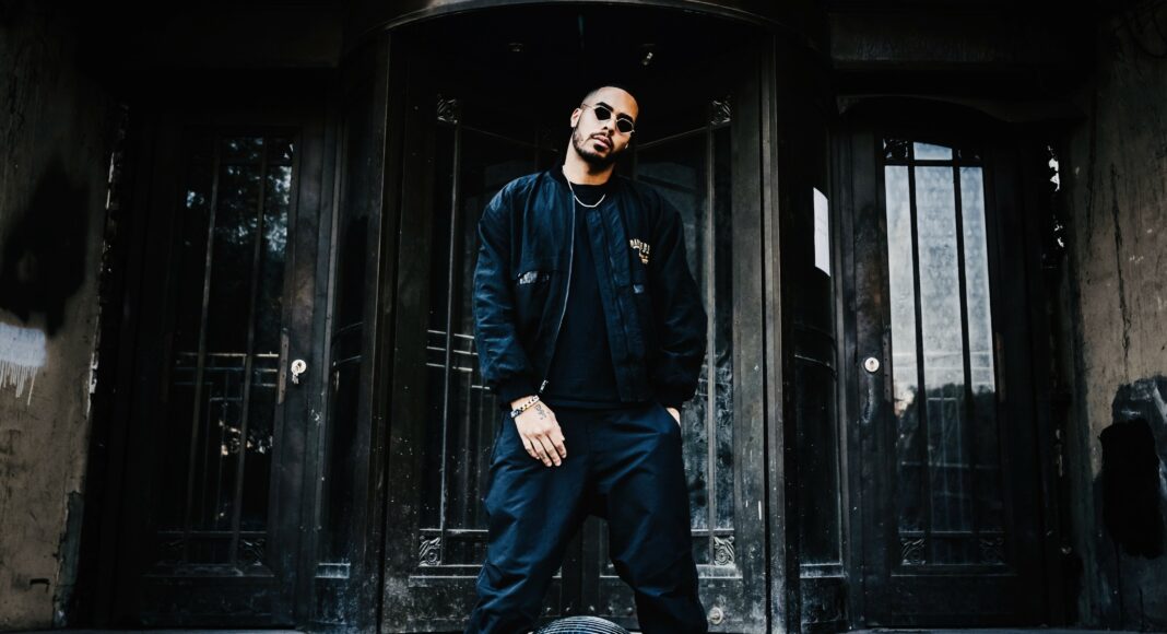 A man in dark clothing and sunglasses stands on stone steps in front of a large doorway, with a silver disco ball at his feet.