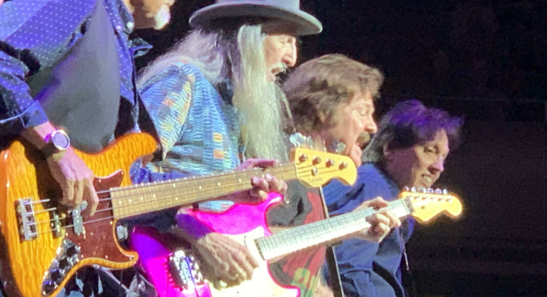 Four musicians play electric guitars on stage under bright lights. The man in front has a long white beard and wears a wide-brimmed hat. The focus is on their hands and instruments as they perform energetically.