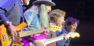 Four musicians play electric guitars on stage under bright lights. The man in front has a long white beard and wears a wide-brimmed hat. The focus is on their hands and instruments as they perform energetically.