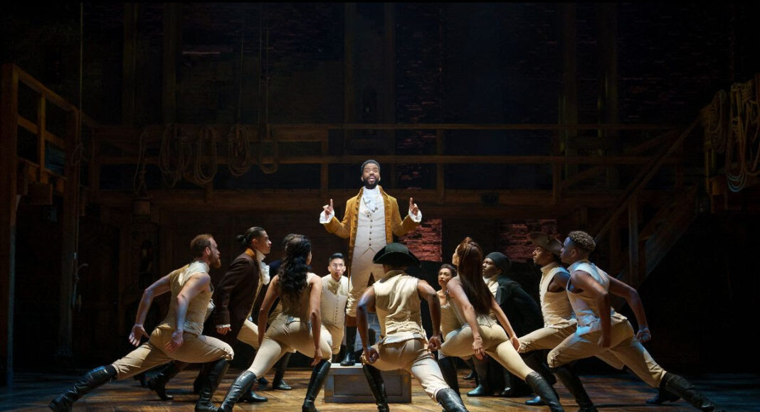 A group of performers in historical costumes dance on stage, centered around a man standing on a table with his finger raised, under dramatic stage lighting and a wooden set backdrop.