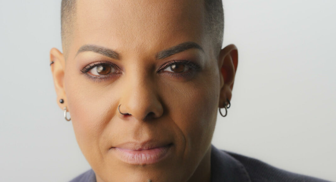 A person with short hair, wearing small hoop earrings and a nose ring, looks confidently into the camera against a plain background. They are wearing a collared, textured blazer.