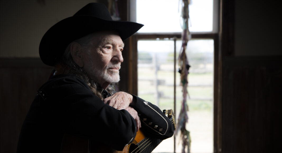 An older man in a black cowboy hat and black jacket sits indoors, resting his arms on an acoustic guitar, looking thoughtfully out a window with soft daylight streaming in.