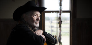 An older man in a black cowboy hat and black jacket sits indoors, resting his arms on an acoustic guitar, looking thoughtfully out a window with soft daylight streaming in.