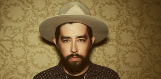 Bearded man wearing a hat and standing against a wallpaper background