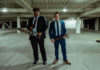 Two men in suits in a garage. The black man has a chainsaw; the white man has a hatchet