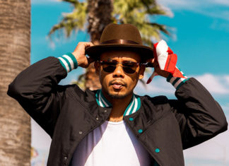 Man in sunglasses, his hands touching his hat, standing by palm trees