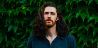 Pale man with dark long hair and beard in front of greenery