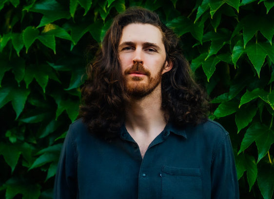 Pale man with dark long hair and beard in front of greenery