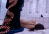 Woman with stitches along the length of her spine crumpled on a bathroom floor with a caped figure looming over her