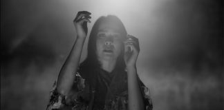 Young woman in a video still, raising her hands up in a dramatic manner