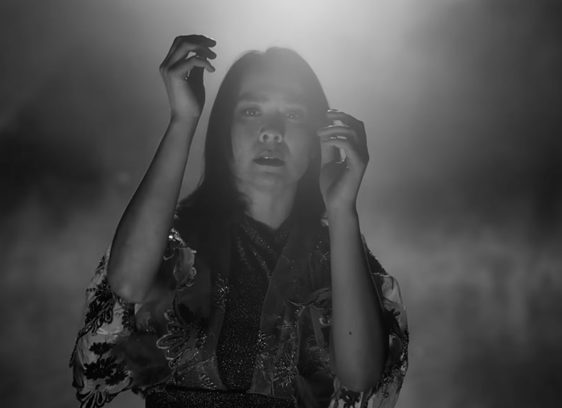 Young woman in a video still, raising her hands up in a dramatic manner