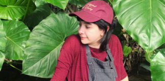 Person dressed in red shirt and hat in overalls in front of a lush green plant