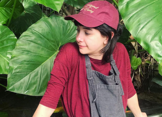 Person dressed in red shirt and hat in overalls in front of a lush green plant