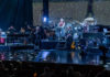 Large ensemble of musicians playing on a stage with holiday lights