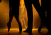 Couple in 1920s clothing seen behind silhouettes of dancers' legs on a dramatically lit stage