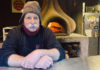 Man wearing a cap sitting in front of a wood-fired oven