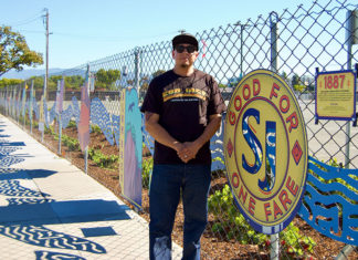Man standing next to a chainlink fence with a piece of art hanging on i